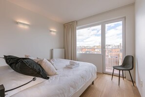 A hotel room with a bed, a chair, a window with a view of buildings, and a ceiling light. - Trendy apartment close to the coast (Oostende)
