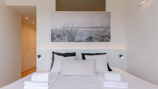 A neatly made bed with white linens and pillows, a painting of a beach scene above it, and a door leading to another room. - Trendy apartment close to the coast (Oostende)