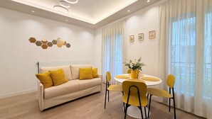 Un salon moderne avec un canapé beige, des coussins jaunes, une table de salle à manger ronde avec des chaises jaunes et une grande fenêtre ornée de rideaux légers.