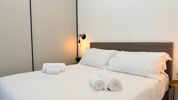 Un lit bien fait, avec du linge de lit blanc, une tête de lit et un luminaire fixé au mur.