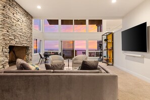 Ein Wohnzimmer mit Kamin aus Stein, einem großen Fenster mit Sonnenuntergangsblick, einem Flachbildfernseher und einem Bücherregal.
