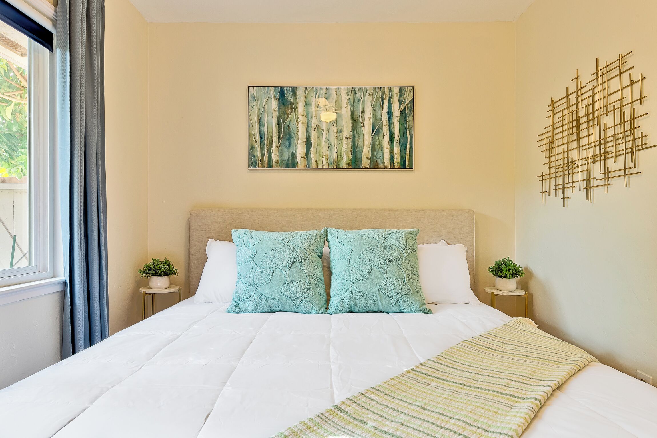 A neatly made bed with a white comforter and two matching pillows, a beige headboard, a wall-mounted abstract art piece, and a small potted plant on a bedside table.
