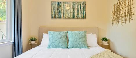 A neatly made bed with a white comforter and two matching pillows, a beige headboard, a wall-mounted abstract art piece, and a small potted plant on a bedside table.
