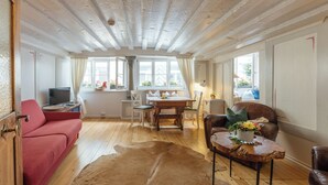 A room with a wooden ceiling, a sofa, a dining table with chairs, a leather armchair, and a coffee table with flowers. - Listed 2-room apartment with historic Lindau ceiling (Lindau)