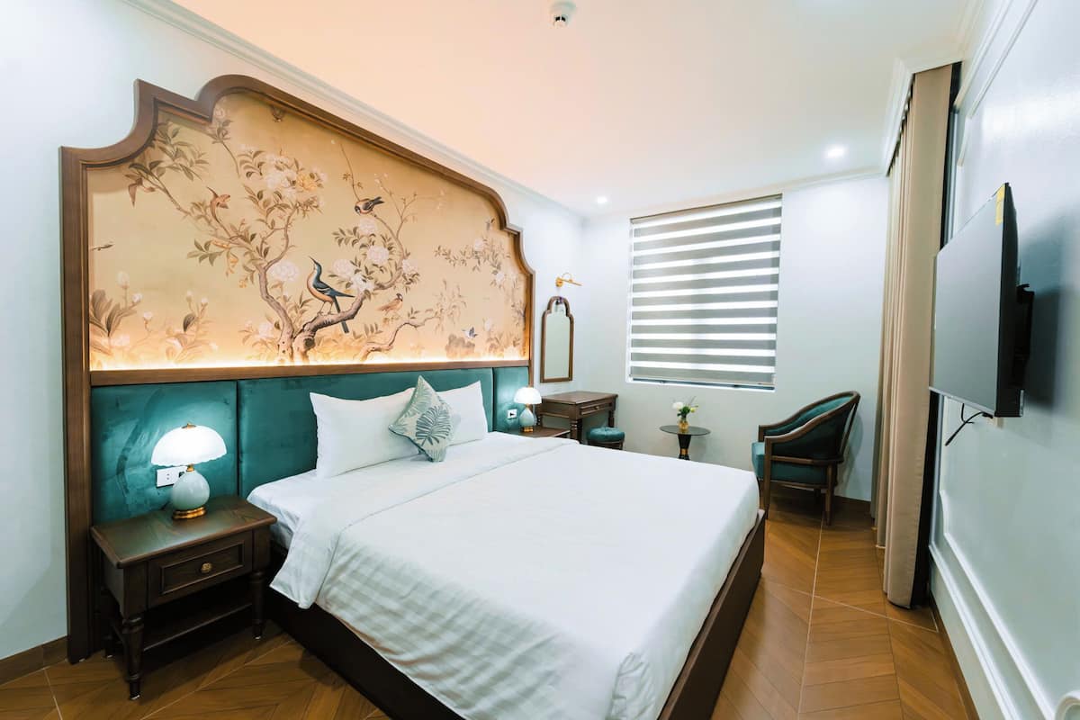 Un dormitorio ordenado con cama, mesitas de noche, lámpara, silla, televisión y ventana con persianas.