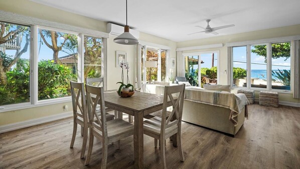 A spacious room with a dining table and chairs, a sofa, and large windows offering a view of the outdoors.