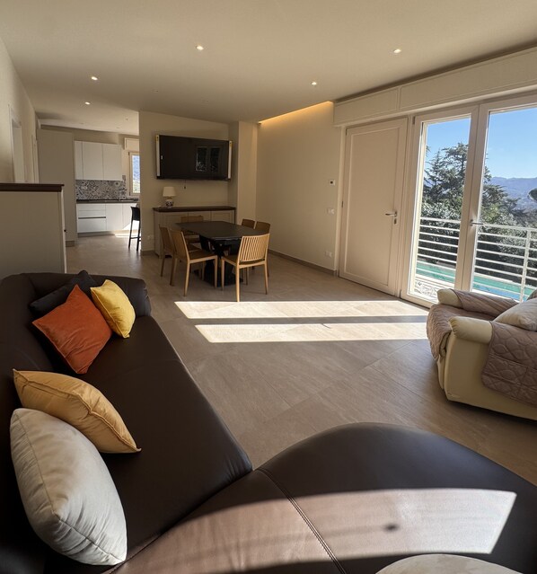 A modern living room with a sofa, a dining table with chairs, a kitchen area, and a balcony with a view.