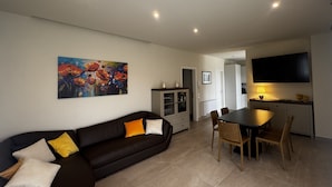 Un salon avec un canapé noir, une table à manger avec des chaises, un coin cuisine avec un téléviseur et un tableau accroché au mur.