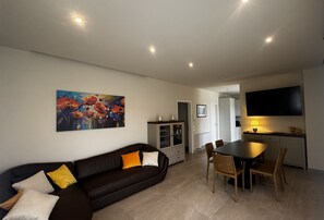 A living room with a black sofa, a dining table with chairs, a kitchen area with a TV, and a painting on the wall.