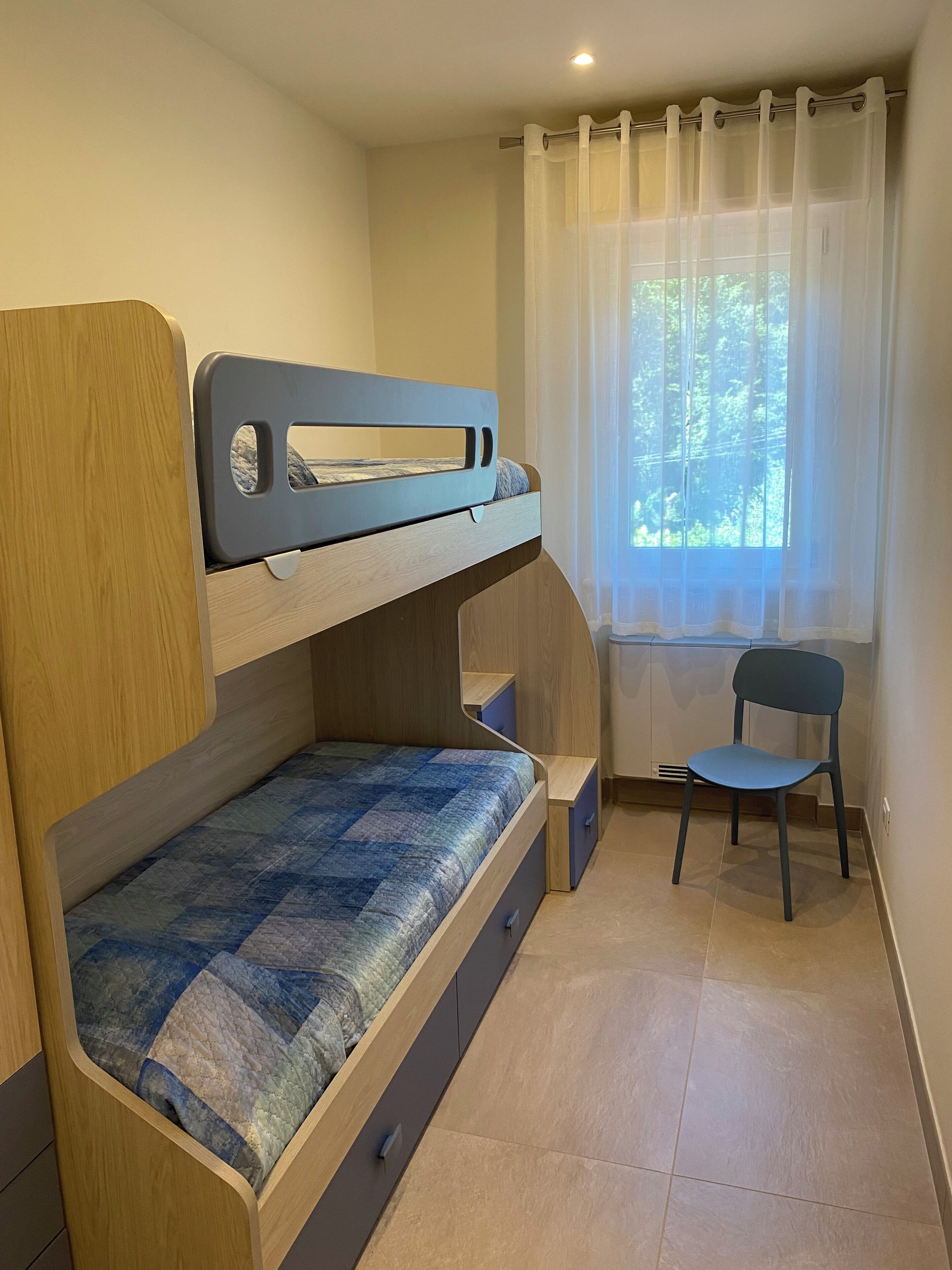 Ein Stockbett mit Schreibtisch und Stuhl, ein Fenster mit Vorhängen und ein gefliester Boden.