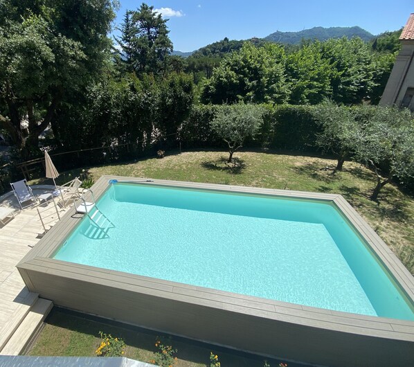 A rectangular swimming pool with a wooden deck surrounding it, set in a backyard with a view of a mountain range.
