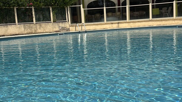 En swimmingpool med krystalklart vand, en betonbygning med balkoner og en solrig himmel.