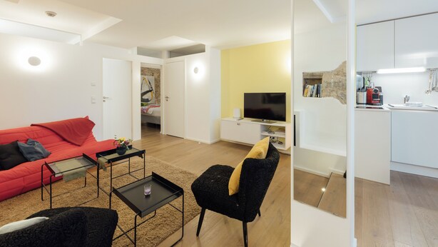 Un salon moderne avec un canapé rouge, un fauteuil noir et un téléviseur. On y trouve également un coin cuisine avec des meubles blancs et un mur jaune.