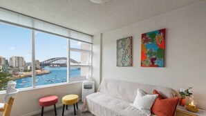Een moderne woonkamer met een groot raam met uitzicht op een haven, een witte bank, kleurrijke krukken en abstracte kunst aan de muur.