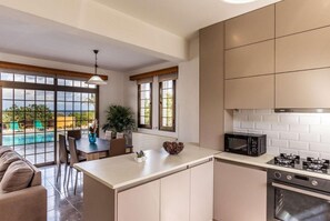 A modern kitchen with a central island, a dining area with a view of a pool, and a large window. - Villa Adonis Large Private Pool, Walk to Beach (Argaka)