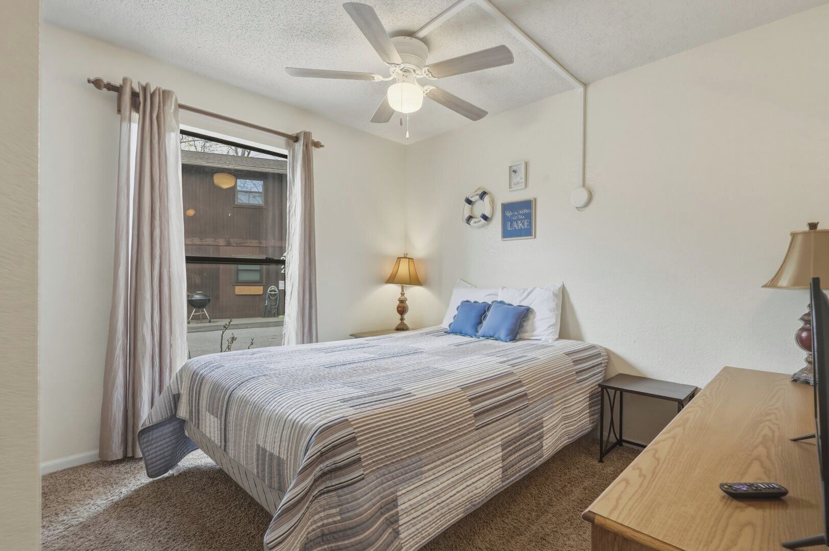 Un dormitorio con cama, ventilador de techo, ventana con cortinas, lámpara de noche y un cuadro colgado en la pared.