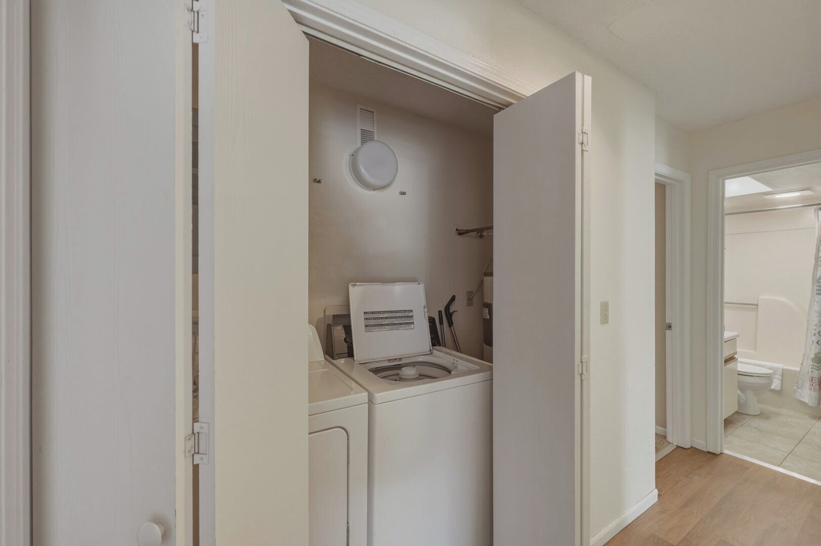 A laundry area with a washing machine and dryer, a ceiling vent, and a door leading to a bathroom with a toilet and shower curtain.