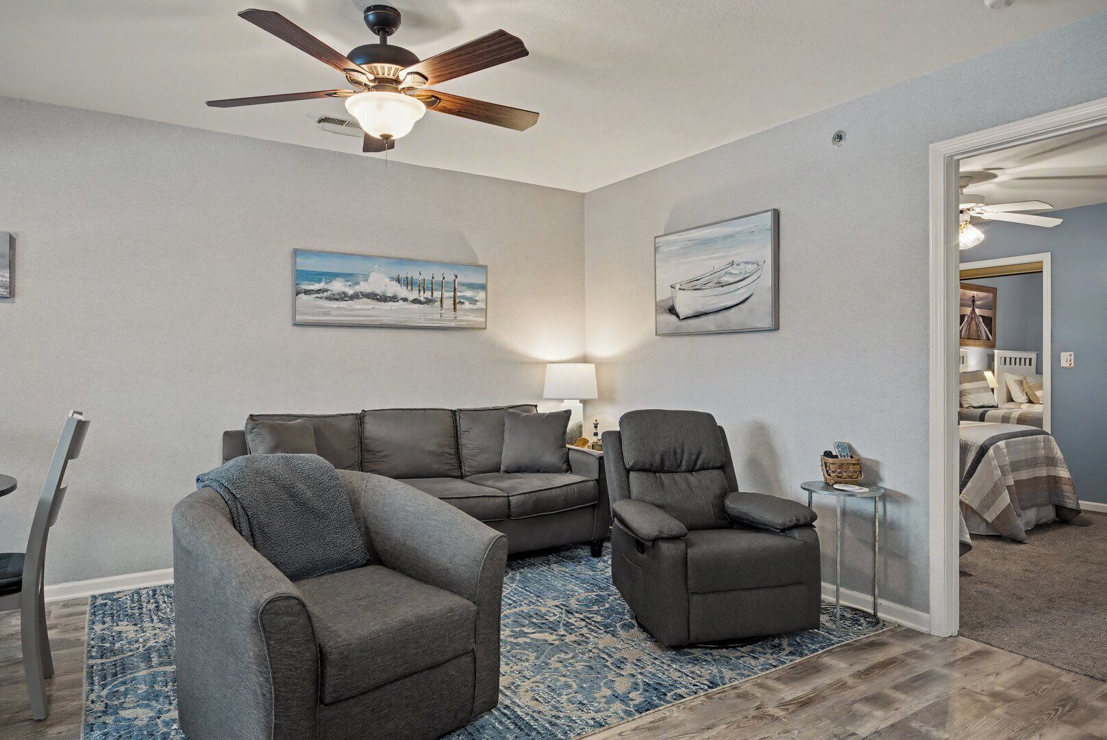 Ein Wohnzimmer mit Deckenventilator, einem grauen Sofa, einem grauen Sessel und einem blauen Teppich mit Muster.