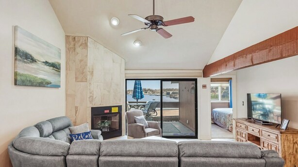 Una sala de estar con un sofá seccional gris, un televisor, un ventilador de techo, una chimenea y un balcón con vistas al agua.
