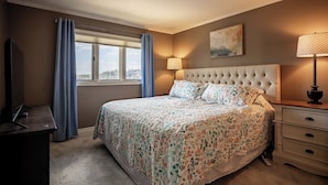 Un dormitorio con cama, televisión, ventana con cortinas, lámpara de noche y un cuadro en la pared.