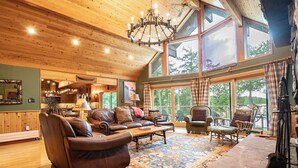 A spacious living room with leather sofas, a coffee table, and a large rug. There are multiple windows providing natural light and views of the outdoors. - Lockout -Reduced Bedrooms - Four Season, Lakefront, Sandy Beach, Amazing Sunsets (Saranac Lake)