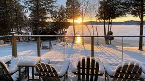 A deck with Adirondack chairs and a view of a sunset over a frozen lake, surrounded by trees. - Lockout -Reduced Bedrooms - Four Season, Lakefront, Sandy Beach, Amazing Sunsets (Saranac Lake)