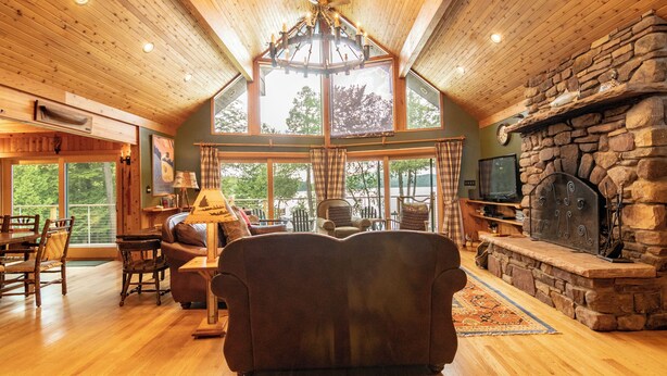 A spacious living room with a stone fireplace, wooden ceiling, and large windows offering a view of the outdoors. - Lockout -Reduced Bedrooms - Four Season, Lakefront, Sandy Beach, Amazing Sunsets (Saranac Lake)