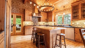 A spacious kitchen with wooden cabinets, a large island with stools, a stone accent wall, and a chandelier. - Lockout -Reduced Bedrooms - Four Season, Lakefront, Sandy Beach, Amazing Sunsets (Saranac Lake)