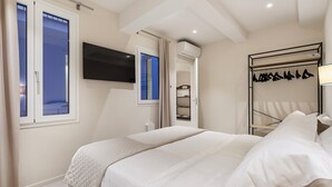 A hotel room with a bed, a TV mounted on the wall, a wardrobe with hanging clothes, and a window with curtains. - Palazzo Accademia-Beato Angelico-2 (Firenze)