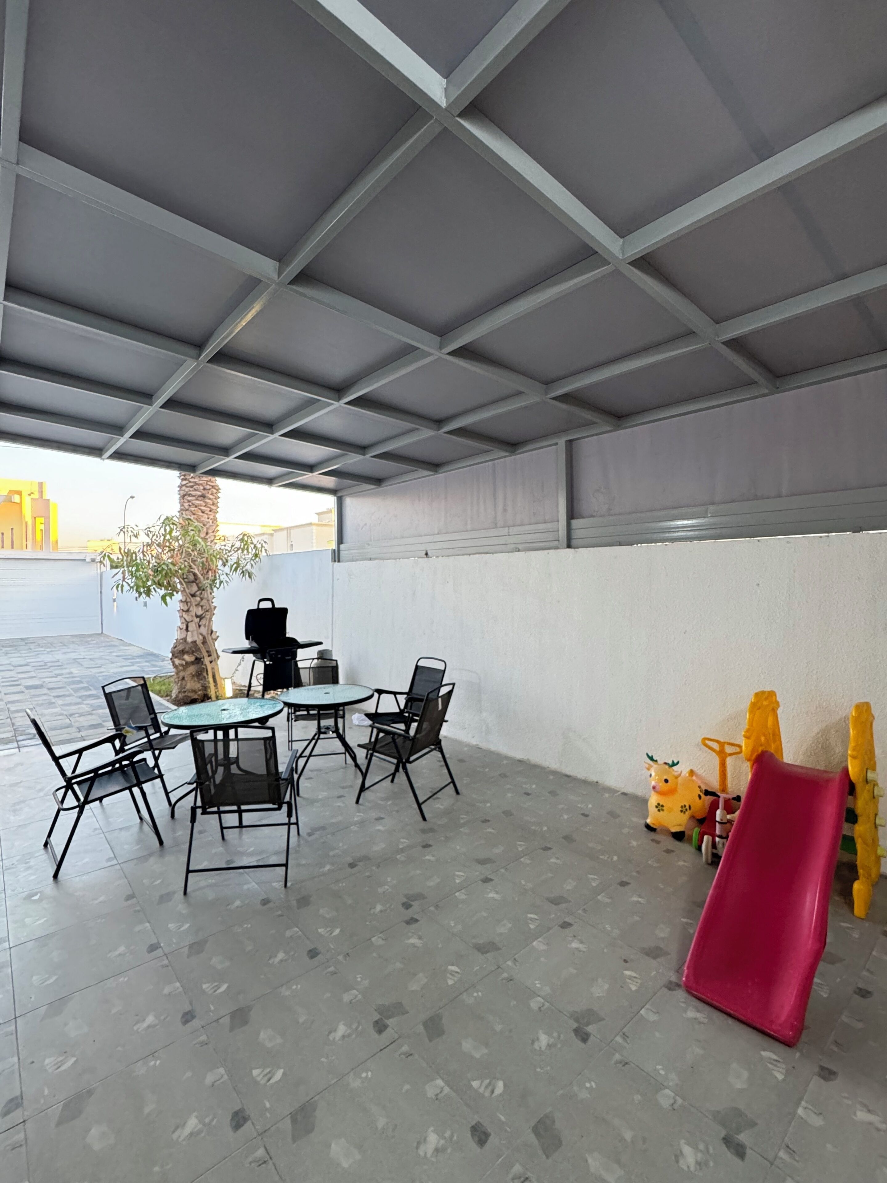 An outdoor patio with a table and chairs, a grill, a palm tree, and a children's play area with a slide.