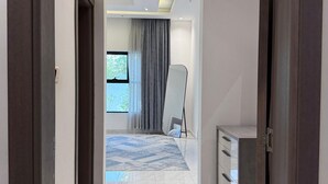 Corredor que dá para um quarto com uma janela grande, uma cama, uma mesa de cabeceira e um guarda-roupa.