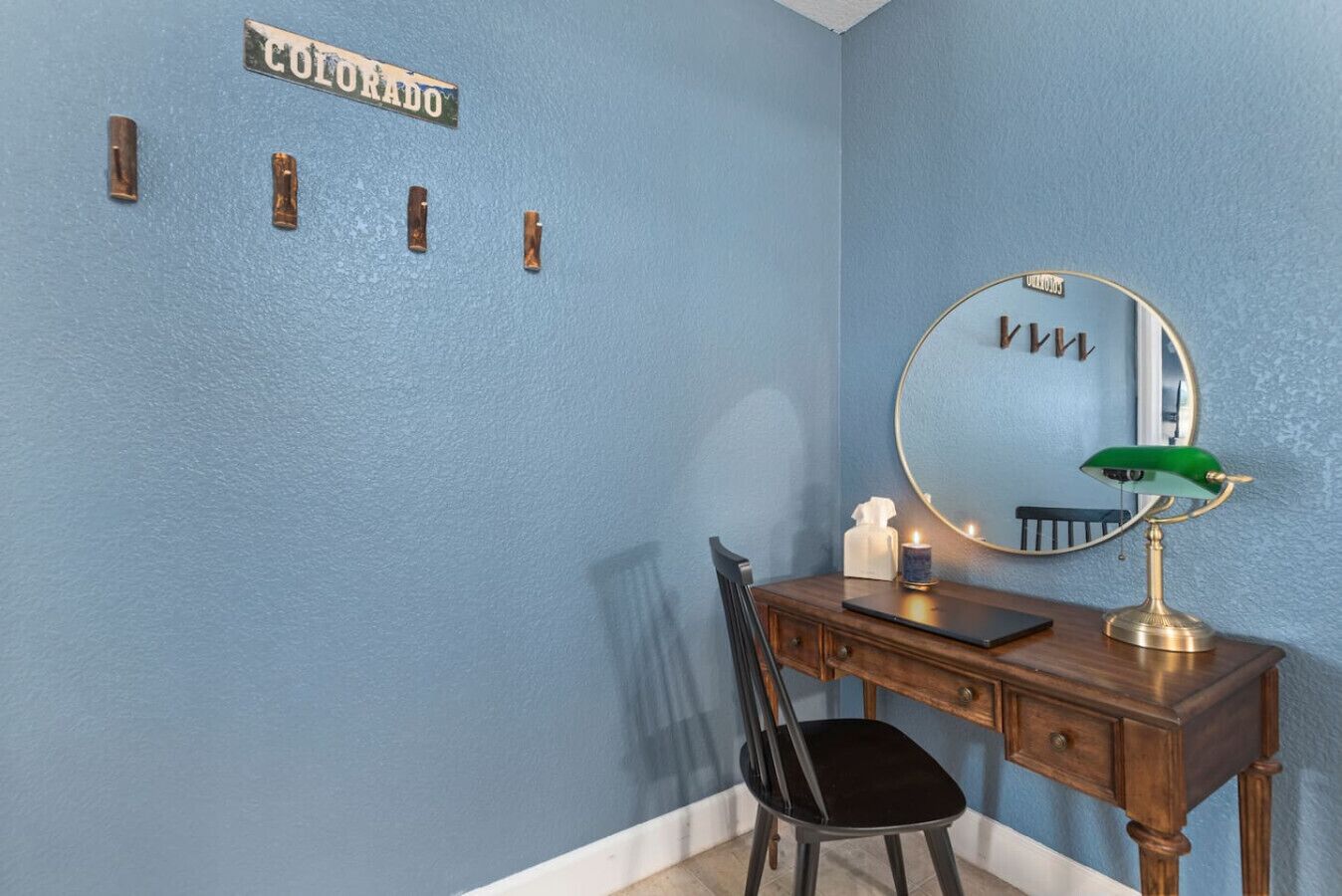 Ein Zimmer mit blauer Wand, einem hölzernen Schminktisch, einem schwarzen Stuhl, einem runden Spiegel und einer grünen Lampe, sowie einem Schild mit der Aufschrift 'COLORADO'.
