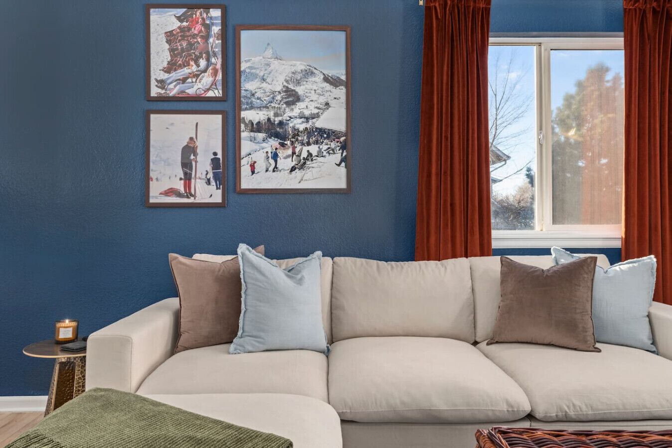 A living room with a beige sofa, three framed pictures on the wall, a window with red curtains, and a small side table with a candle.