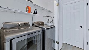 Un lavadero con dos lavadoras y secadoras Samsung de carga frontal, un estante con productos de limpieza y una puerta blanca.