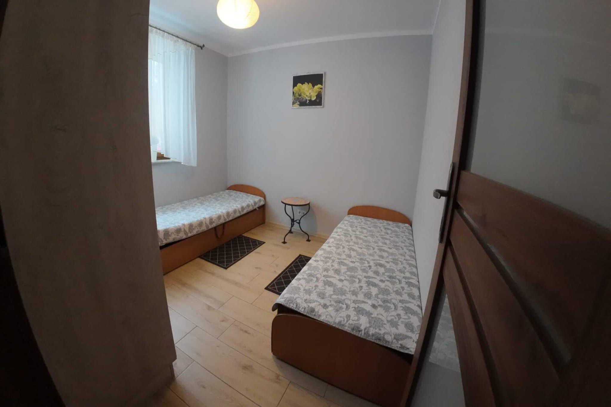 A small room with two beds, a window with curtains, a small table, and a wall-mounted picture.