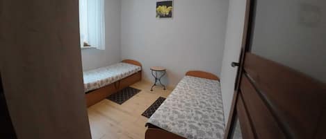 Una stanza modesta con due letti, una finestra con tende, un tavolino e un quadro appeso al muro.