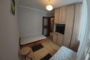 A small, compact room with a bed, a small table, a cabinet, and a door leading to a kitchen area.