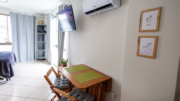 A room with a wooden dining table, chairs, a flat-screen TV, an air conditioner, and framed artwork on the wall. - Loft equipado a poucos passos da praia (Arraial do Cabo)