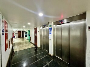 Ένας διάδρομος ξενοδοχείου με ανελκυστήρες, έναν πυροσβεστήρα και μια πινακίδα που υποδεικνύει έναν χώρο κοινωνικής συναναστροφής.