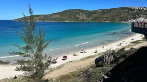 A beach with clear blue water, sandy shore, and a hillside in the background. - Loft equipado a poucos passos da praia (Arraial do Cabo)