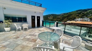 Ein Balkon mit Tisch und Stühlen, einem Blumentopf und Blick auf Strand und Hügel.