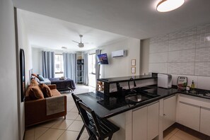 Una cocina moderna con encimera negra, armarios blancos y horno integrado. Hay una zona de comedor con un sofá marrón y una silla negra. Un televisor está montado en la pared, y se ve un ventilador de techo.