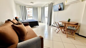 A hotel room with a bed, a TV, a desk with chairs, and a window with curtains. - Loft equipado a poucos passos da praia (Arraial do Cabo)