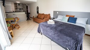A bedroom with a bed, a sofa, a dining table with chairs, a kitchen area, and a ceiling fan. - Loft equipado a poucos passos da praia (Arraial do Cabo)