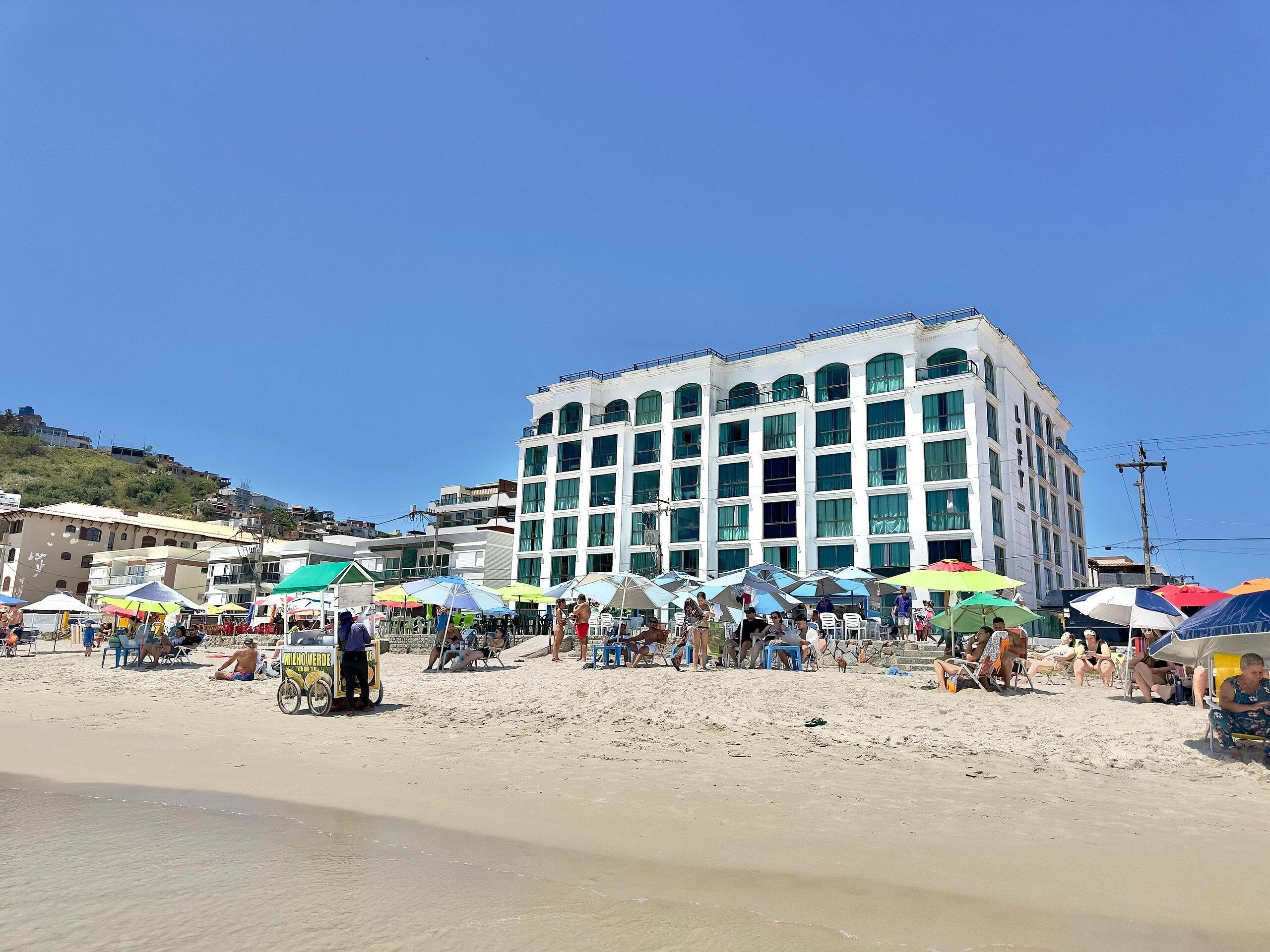 Un edificio de varios pisos con grandes ventanales y techo plano, ubicado en una playa arenosa con sombrillas y sillas de playa.
