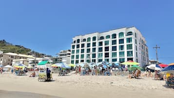 Un immeuble à plusieurs étages, doté de grandes fenêtres et d’un toit plat, situé sur une plage de sable, avec des parasols et des chaises de plage.
