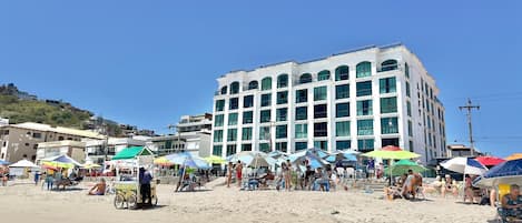A multi-story building with large windows and a flat roof, situated on a sandy beach with umbrellas and beach chairs.