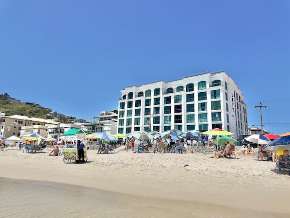 A multi-story building with large windows and a flat roof, situated on a sandy beach with umbrellas and beach chairs. - Loft equipado a poucos passos da praia (Arraial do Cabo)