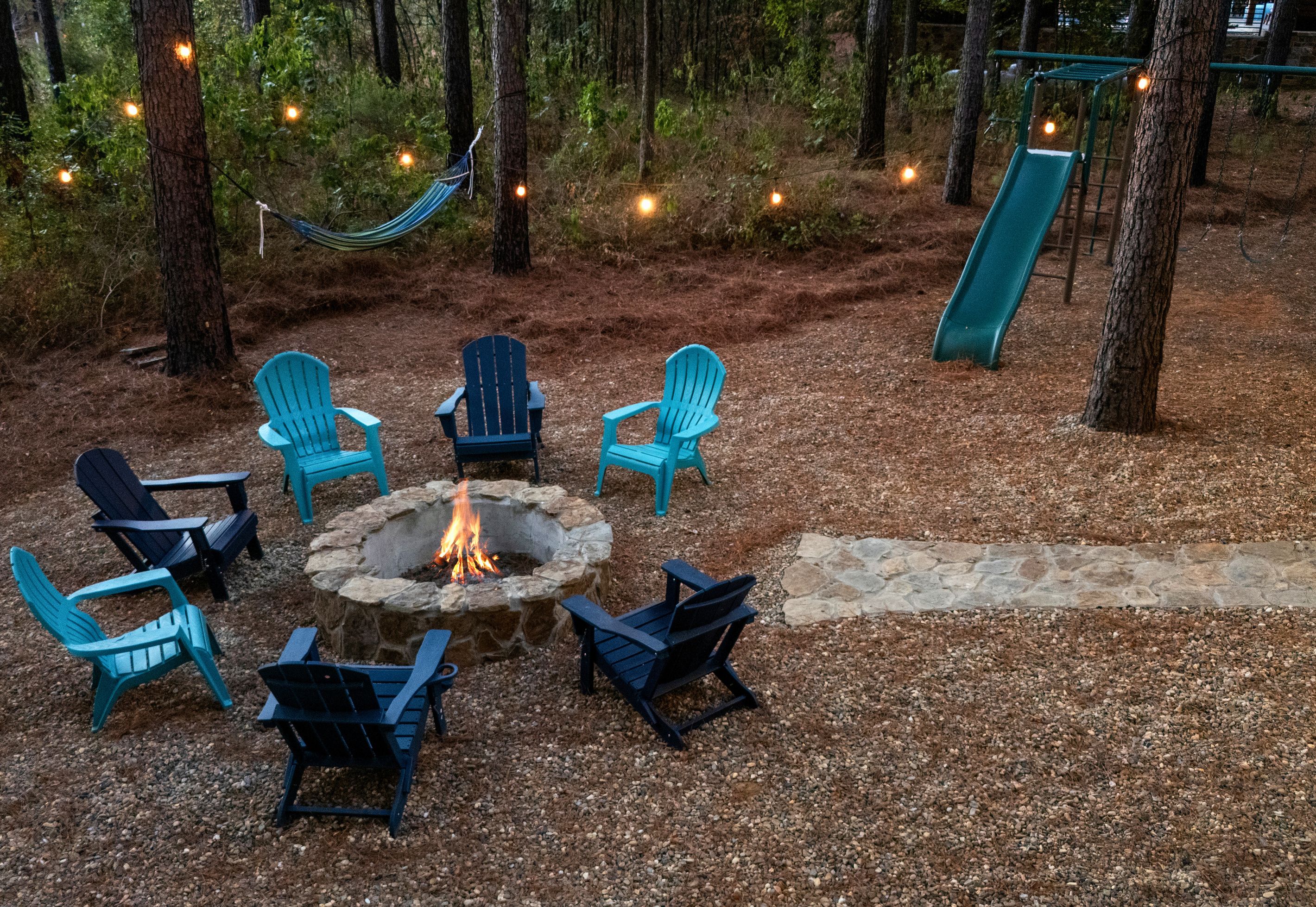 An outdoor seating area with chairs arranged around a stone fire pit, surrounded by trees and a hammock.