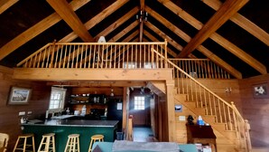 Um loft espaçoso com teto alto, vigas de madeira, um espaço bar e uma área de estar aconchegante com sofá e banquetas.